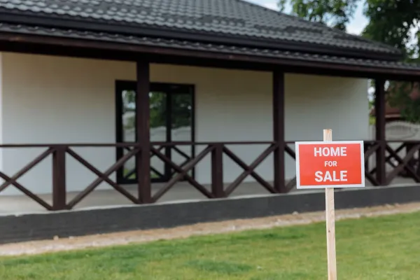 A red home-for-sale sign staked in a front lawn in front of a house with a covered porch