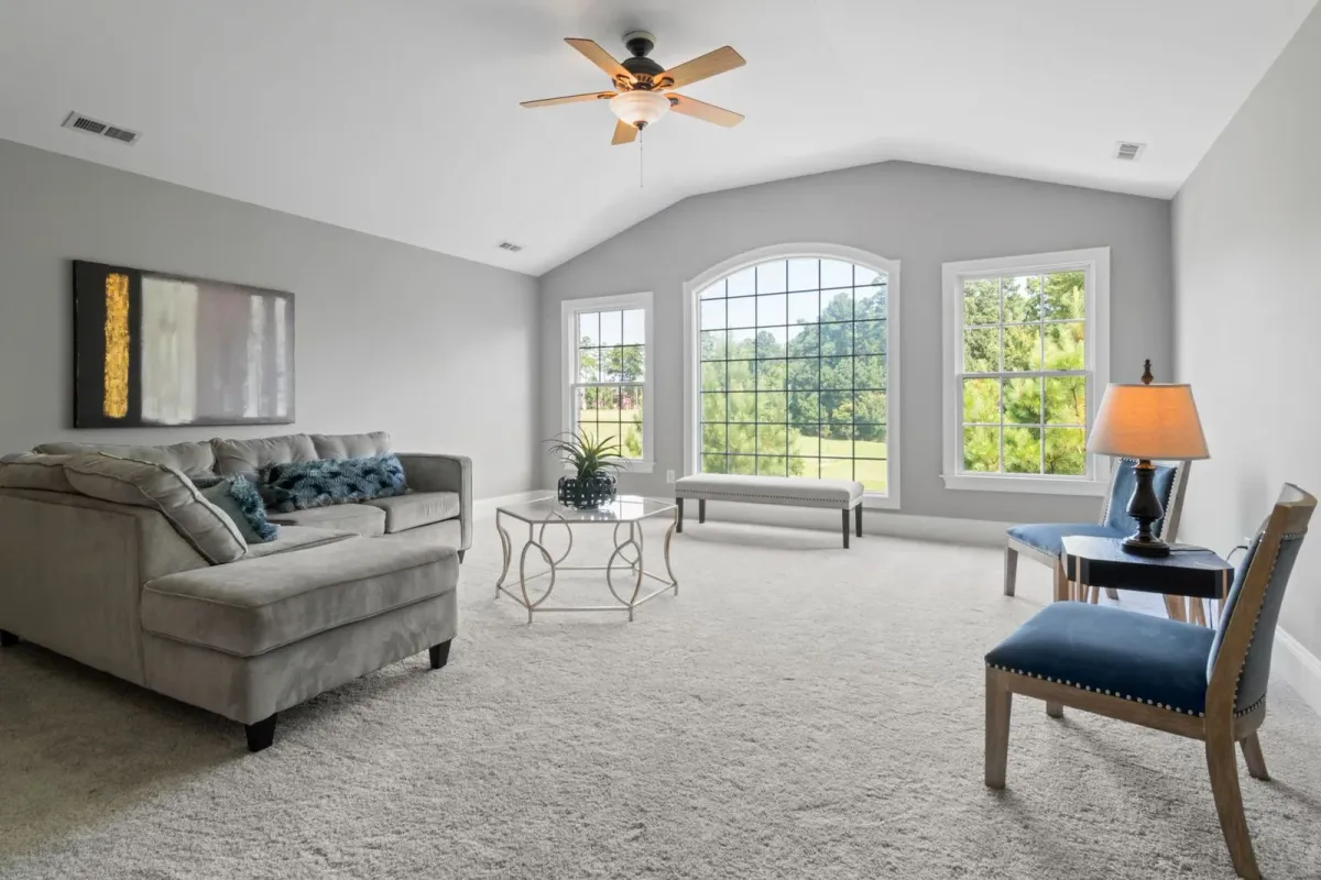 A staged living room with a gray sectional, a glass coffee table, and a large arched window under a vaulted ceiling